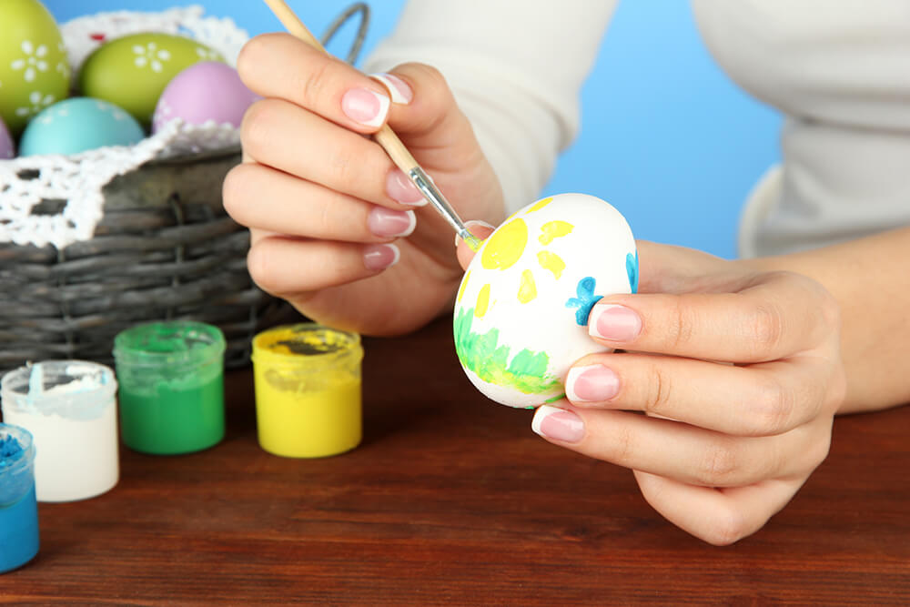 Young woman painting Easter eggs on color background